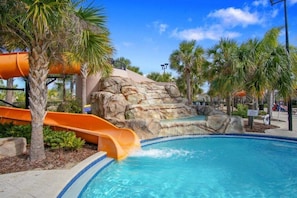 A pool area with a water slide, palm trees, and a rocky backdrop.