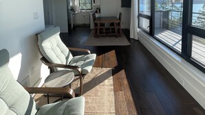 Una sala moderna con sofá, sillas y una mesa de comedor. Se observa una cocina al fondo y ventanas amplias con vistas al exterior.