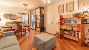 A cozy living room with a wooden floor, a grey sofa, a dining table with chairs, a bookshelf filled with books, and a mirror on the wall. - 5' from Bocconi University with 2 Bedr and 2 Bathr (Milano)