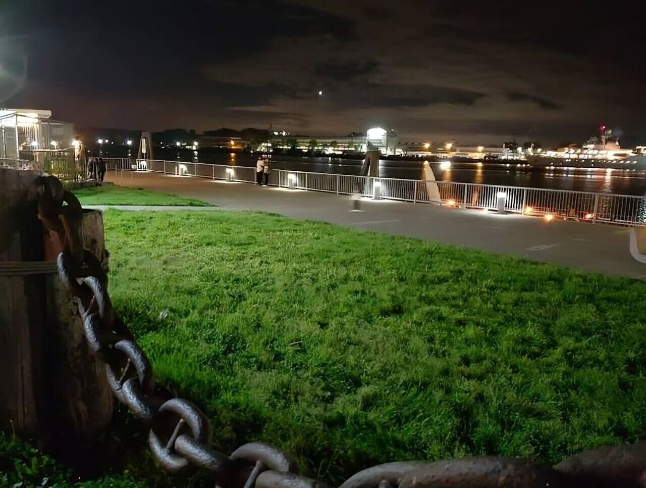 A nighttime view of a riverside walkway with a grassy area and a metal fence.