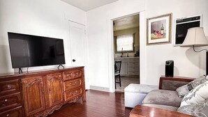 Ein Wohnzimmer mit Flachbildfernseher, Holzschrank, Sofa und einem durchgegangene Tür erkennbaren Küchenbereich.