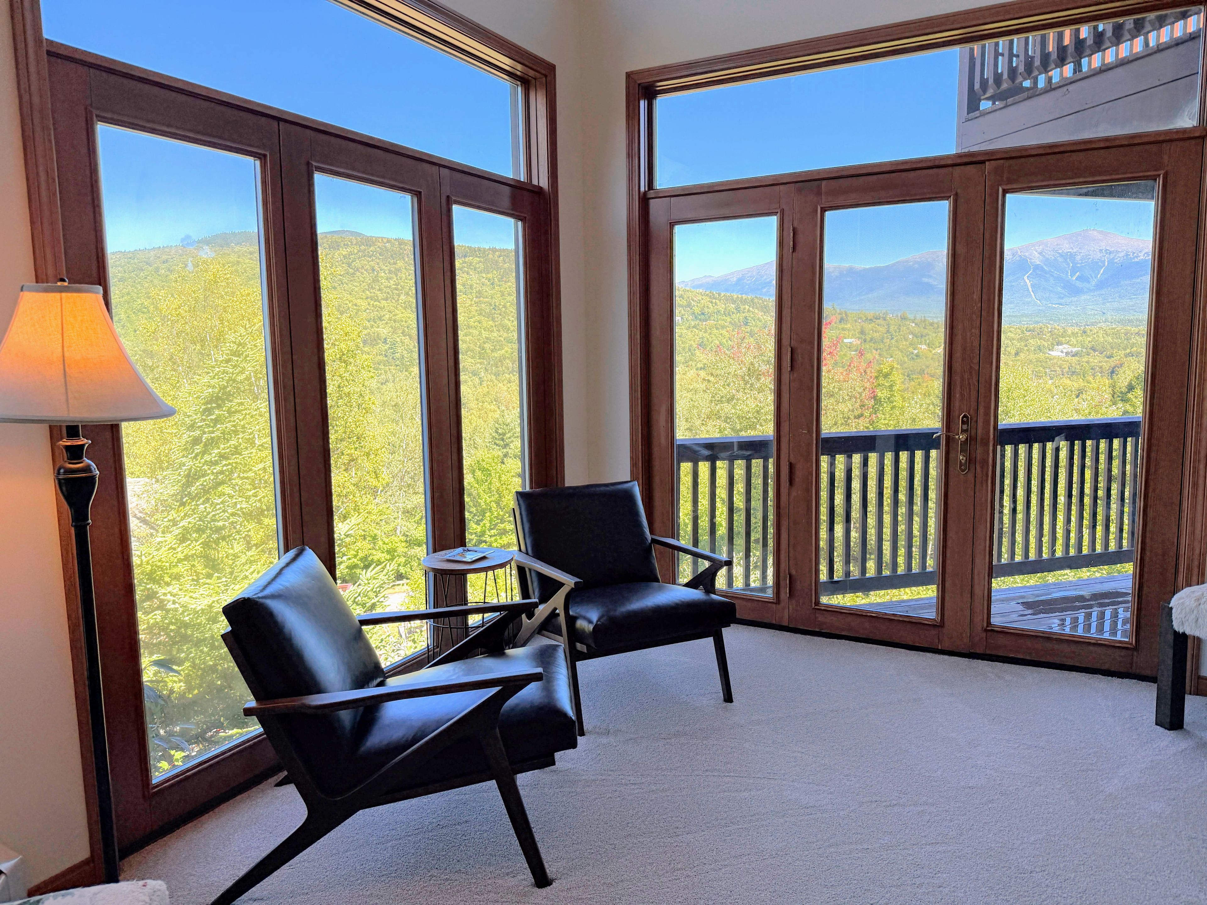 Ein Zimmer mit einer großen Schiebetür, die auf einen Balkon mit Bergblick führt. Zwei schwarze Ledersessel und ein kleiner Tisch mit Lampe stehen dort.
