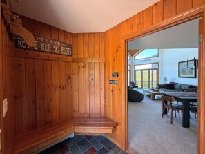 Un intérieur en bois avec un panneau indiquant qu’il faut enlever les chaussures de ski, un banc et un coin repas avec une table et des chaises.