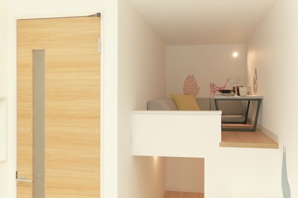 Una habitación moderna y compacta con puerta de madera, una pequeña zona de estar con cojines y una mesa con lámpara.