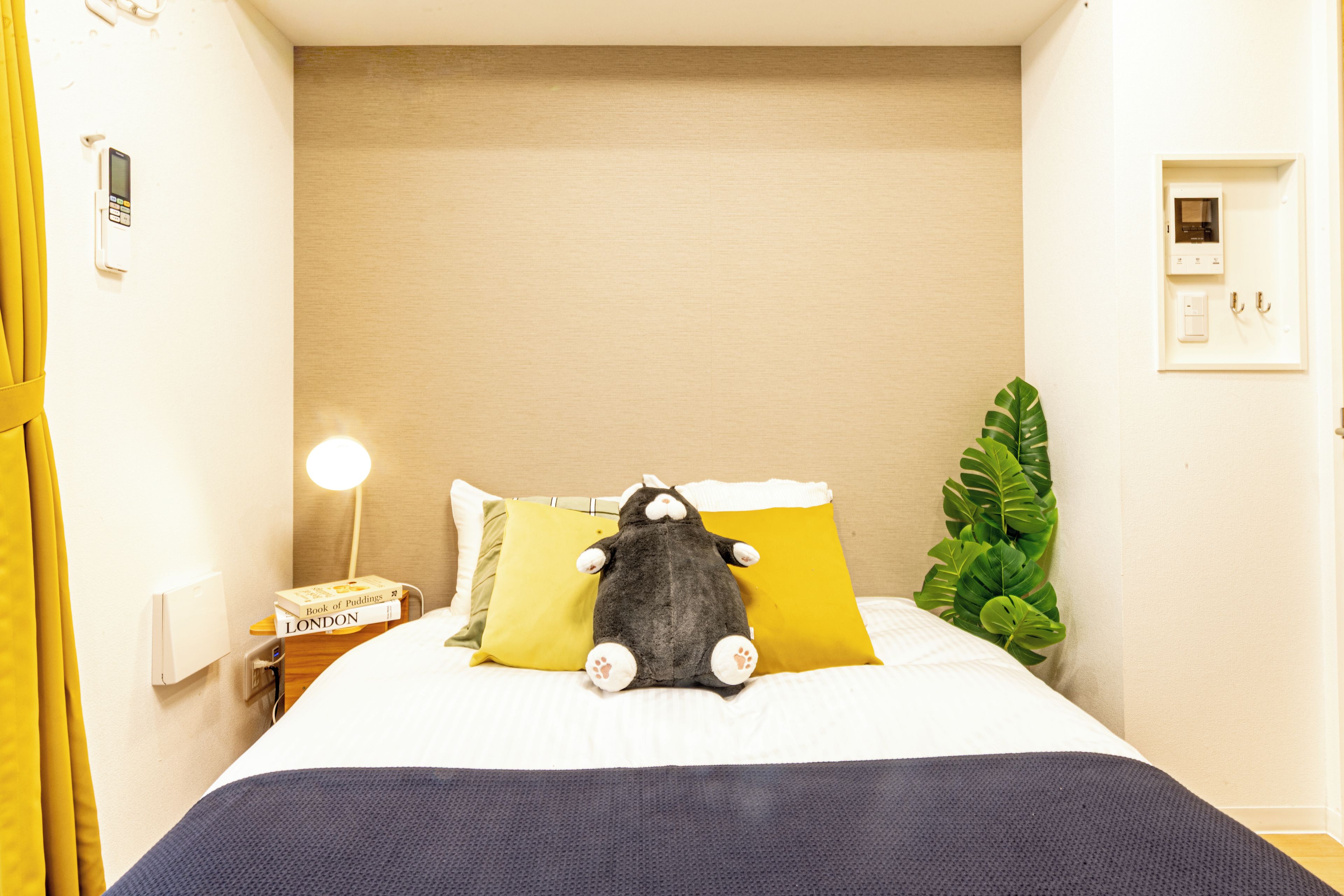 Una cama bien hecha con un peluche gris, una lámpara de noche, un libro y una planta en maceta.