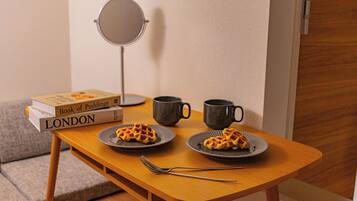 A small wooden table with two plates of waffles, a book titled 'Book of Puddings', a mirror, and two mugs.