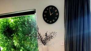 A wooden table with two stools, a wall clock, a vase with branches, and a window with greenery outside. - Whakatā Place - Peaceful, Chic, Central Retreat Near Beaches, Parks & Adventure! (Motueka)