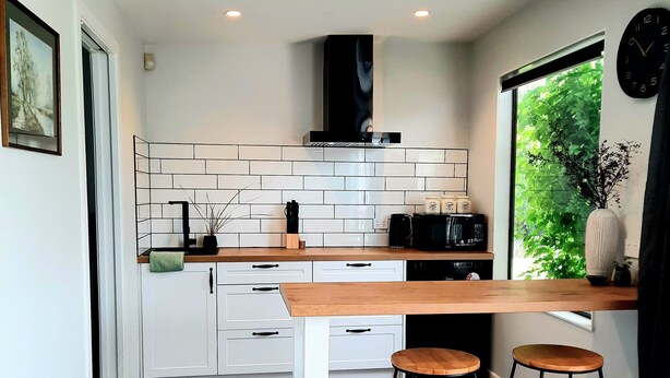 A modern kitchen with white cabinetry, a wooden countertop, two stools, and a window with a view of greenery. - Whakatā Place - Peaceful, Chic, Central Retreat Near Beaches, Parks & Adventure! (Motueka)