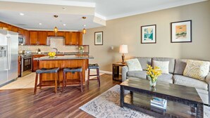 A modern kitchen with wooden cabinets, a central island, and bar stools. There is a dining area with a gray sofa and a coffee table.