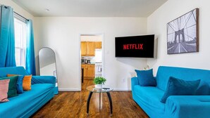 Een woonkamer met twee blauwe banken, een salontafel, een televisie waarop 'NETFLIX' staat en een keuken op de achtergrond.