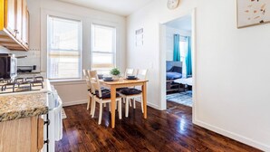 A compact kitchen with a stove, dining table, and chairs. There are two windows with blinds, a clock on the wall, and a door leading to a bedroom. - 2BR Apt| Free Pkng| Netflix|NJ Train| Pets |5 pple (Newark)
