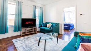 A living room with a television displaying 'NETFLIX', a teal sofa, a glass coffee table, and a blue couch. - 2BR Apt| Free Pkng| Netflix|NJ Train| Pets |5 pple (Newark)