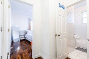 Quarto com cama, mesa de cabeceira e janela. O banheiro possui banheira, vaso sanitário e pia.