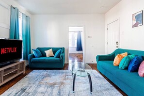 Uma sala de estar com uma TV exibindo 'NETFLIX', um sofá verde-água, uma mesa de centro de vidro e um móvel para TV de madeira.