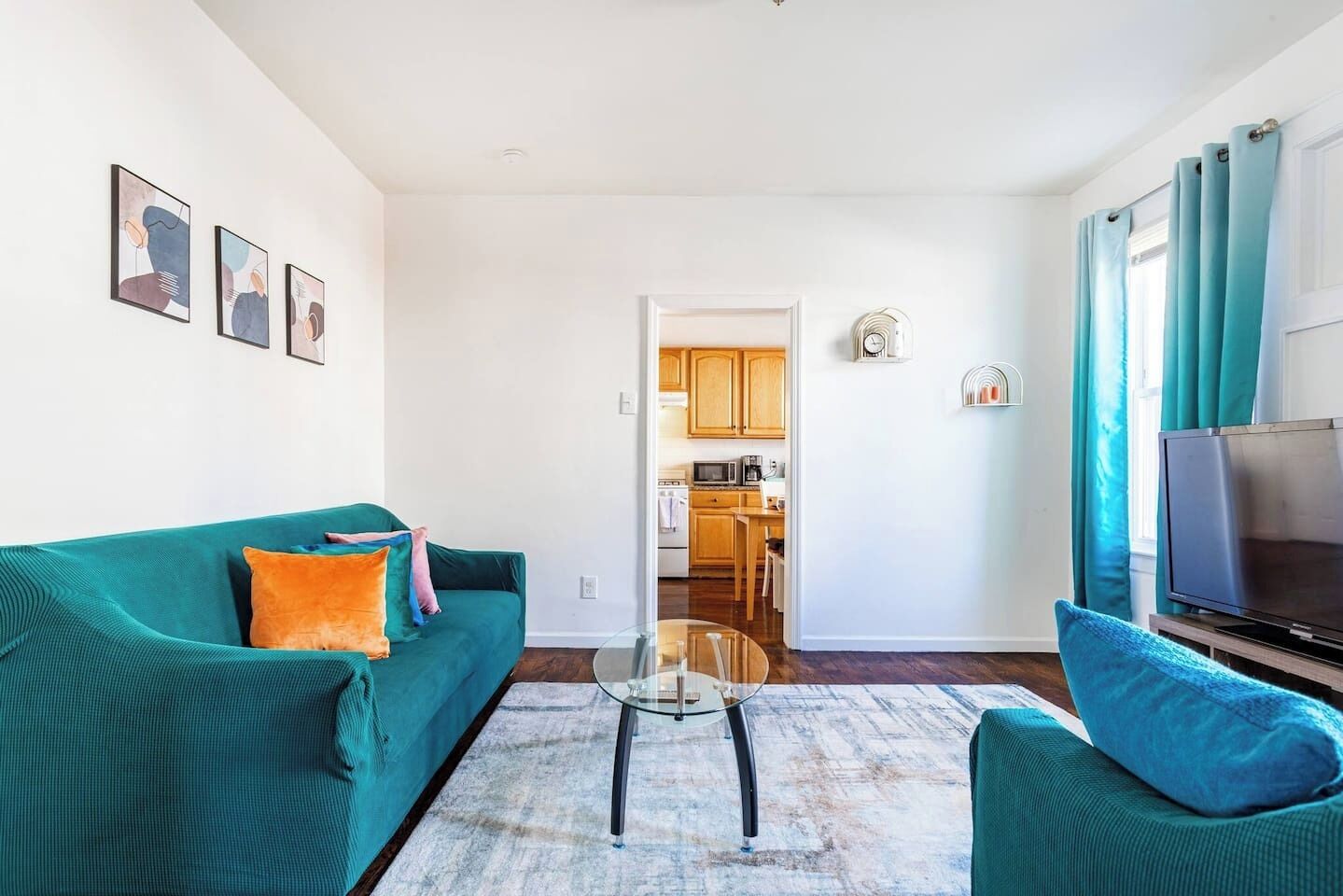 Una sala de estar con sofás color turquesa, una mesa de centro de vidrio y un televisor sobre un mueble. Se ve una cocina al fondo.