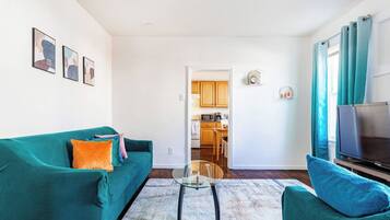 Una sala de estar con sofás color turquesa, una mesa de centro de vidrio y un televisor sobre un mueble. Se ve una cocina al fondo.