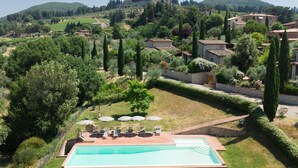 Ein Schwimmbad mit Liegestühlen und Sonnenschirmen, umgeben von einem gepflegten Garten und Blick auf eine hügelige Landschaft mit Gebäuden.