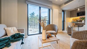 A modern living room with a sofa, a wicker chair, a coffee table, and a view of the outdoors through large windows. - Costa Rogowo Romantyczne Studio Shellter Morze (Trzebiatow)