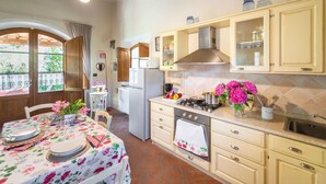 Uma cozinha com armários de madeira, fogão a gás e geladeira. Há uma mesa de jantar com toalha de mesa florida e cadeiras.