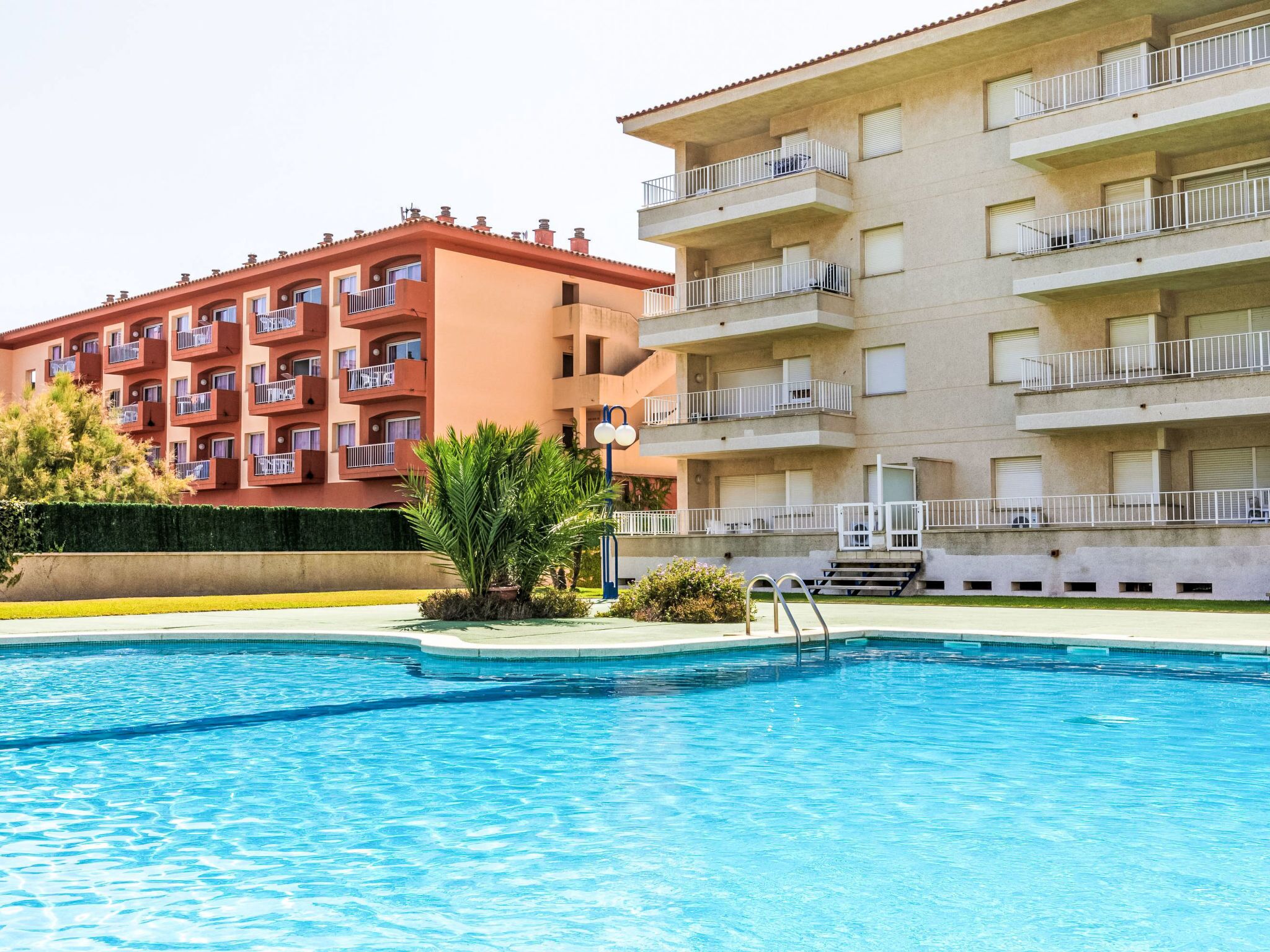 A swimming pool with a clear blue water surface, surrounded by a well-maintained lawn and a hedge. In the background, there are two multi-story buildings with balconies and a red-tiled roof.