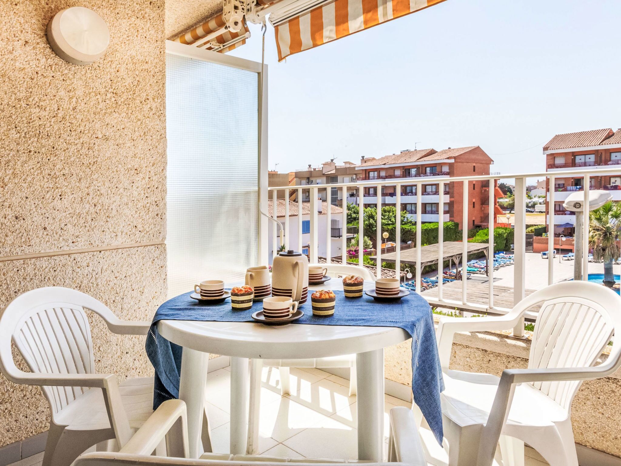 Un balcó amb una taula preparada per esmorzar, caires blancs i vista a una piscina i edificis.