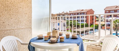 A balcony with a table set for breakfast, white chairs, and a view of a pool and buildings.