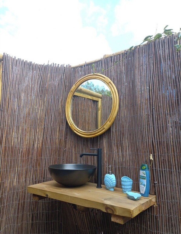 Una cerca de bambú con un espejo redondo, un lavabo negro y un estante de madera con artículos de tocador.