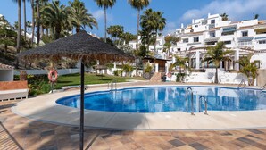 A swimming pool with a thatched umbrella, surrounded by palm trees and white buildings. - Baya de Nerja - Oasis Properties (Nerja)