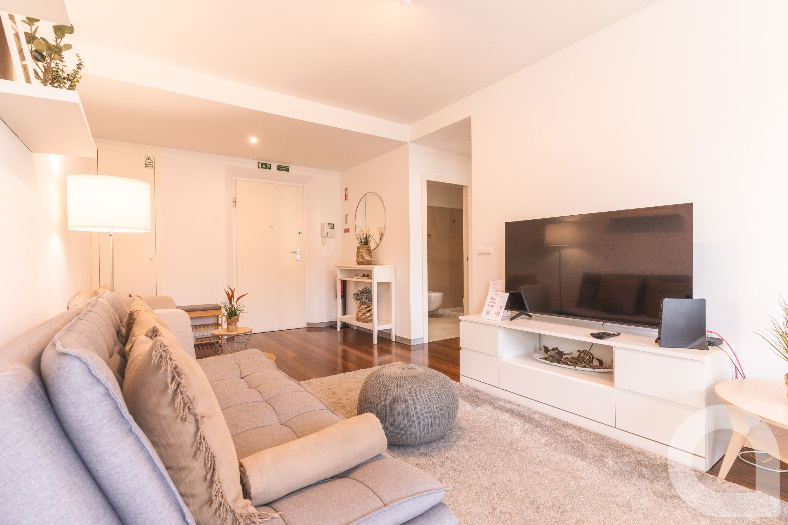 Uma sala de estar moderna com um sofá cinza, uma TV de tela plana e uma mesa pequena com abajur.