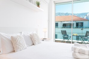 A hotel room with a large bed, white bedding, a view of a building with a red roof, and a balcony with chairs and a table.