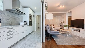 Et moderne køkken med hvide skabe og en marmorbordplade, et opholdsområde med sofa og fjernsyn, samt en trægulv.