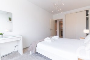 A modern bedroom with a white bed, a bedside table, a desk with a mirror, a hanging light fixture, and a door leading to a bathroom. - Acanto Loft by Atlantic Holiday (Funchal)