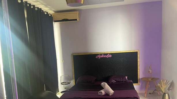 Une chambre à coucher moderne avec un grand lit, une chaise longue noire, un ventilateur de plafond et une petite table d’appoint avec une lampe.