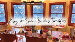 A dining area with a table set for breakfast, a chandelier, and a view of snow-covered trees through large windows. - Wood Nymph Suite at RiverSong Inn in Estes Park - Riverside in Rocky Mountain National Park (Estes Park)