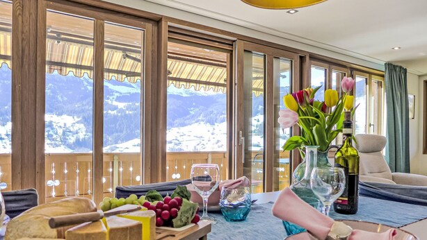 A dining table with a variety of cheeses, fruits, and a bottle of wine, set against a backdrop of a mountain view through large windows. - Chalet Adlerhorst OG by Interhome (Grindelwald)