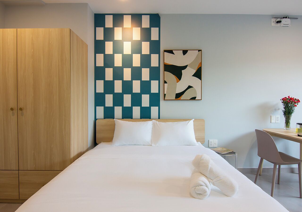 A hotel room with a bed, a desk, a chair, a wardrobe, and a wall with a geometric light fixture.