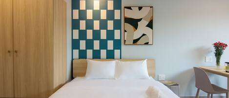 A hotel room with a bed, a desk, a chair, a wardrobe, and a wall with a geometric light fixture.