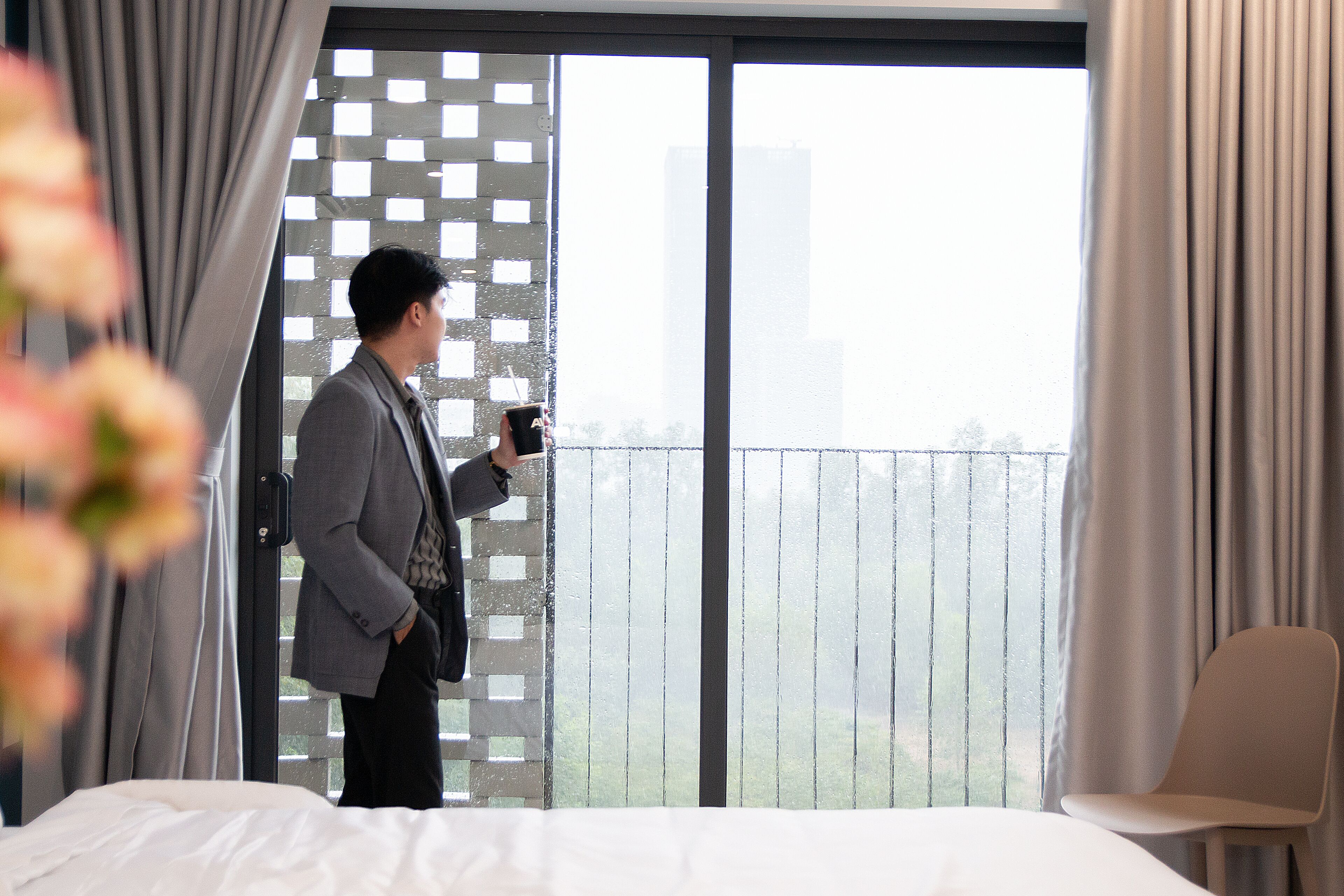A hotel room with a large window, a bed, a chair, and a person holding a phone.