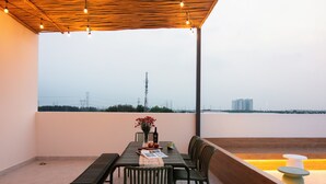 An outdoor dining area with a table and chairs, a view of the city skyline, and a wooden ceiling with hanging lights. - ORA Serviced Apartments - Binh Duong New City (Ho Chi Minh City)