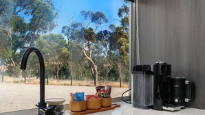 Una cocina con lavamanos, cafeteras y vistas a árboles y una valla.