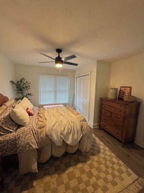 Un dormitorio con cama, cómoda, ventilador de techo, ventana con persianas y una planta.