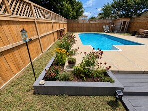 Un patio trasero con piscina, terraza de madera y jardín con flores.