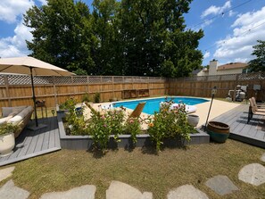 A backyard with a swimming pool, a wooden deck, outdoor furniture, and a fence.