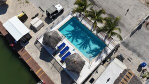 Vista aérea de una zona de piscina con una piscina azul cristalina, sillas de descanso y palmeras.