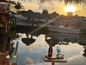 A sunset view with a boat docked at a marina, a table with a bottle and glass, and a building with a sign.