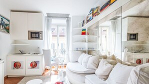 A modern, well-lit living room with a white sofa, a dining area with a table and chairs, a kitchen area with a microwave and cabinets, and a large window with curtains. - Sant' Andrea Fashion Suite by Interhome (Milan)