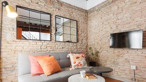 A modern living room with a brick wall, a grey sofa with orange and floral cushions, a round coffee table, and a TV mounted on the wall. - Apartment with a spectacular terrace in the center (Málaga)
