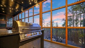 Cozinha externa com churrasqueira de aço inoxidável, janelas amplas e um deck com vista para uma floresta.