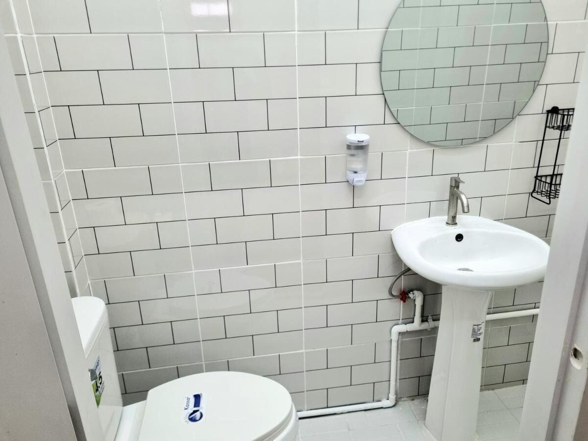 A small, clean bathroom with white tiles, a round mirror, a white sink, and a toilet.