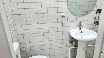 A small, clean bathroom with white tiles, a round mirror, a white sink, and a toilet.